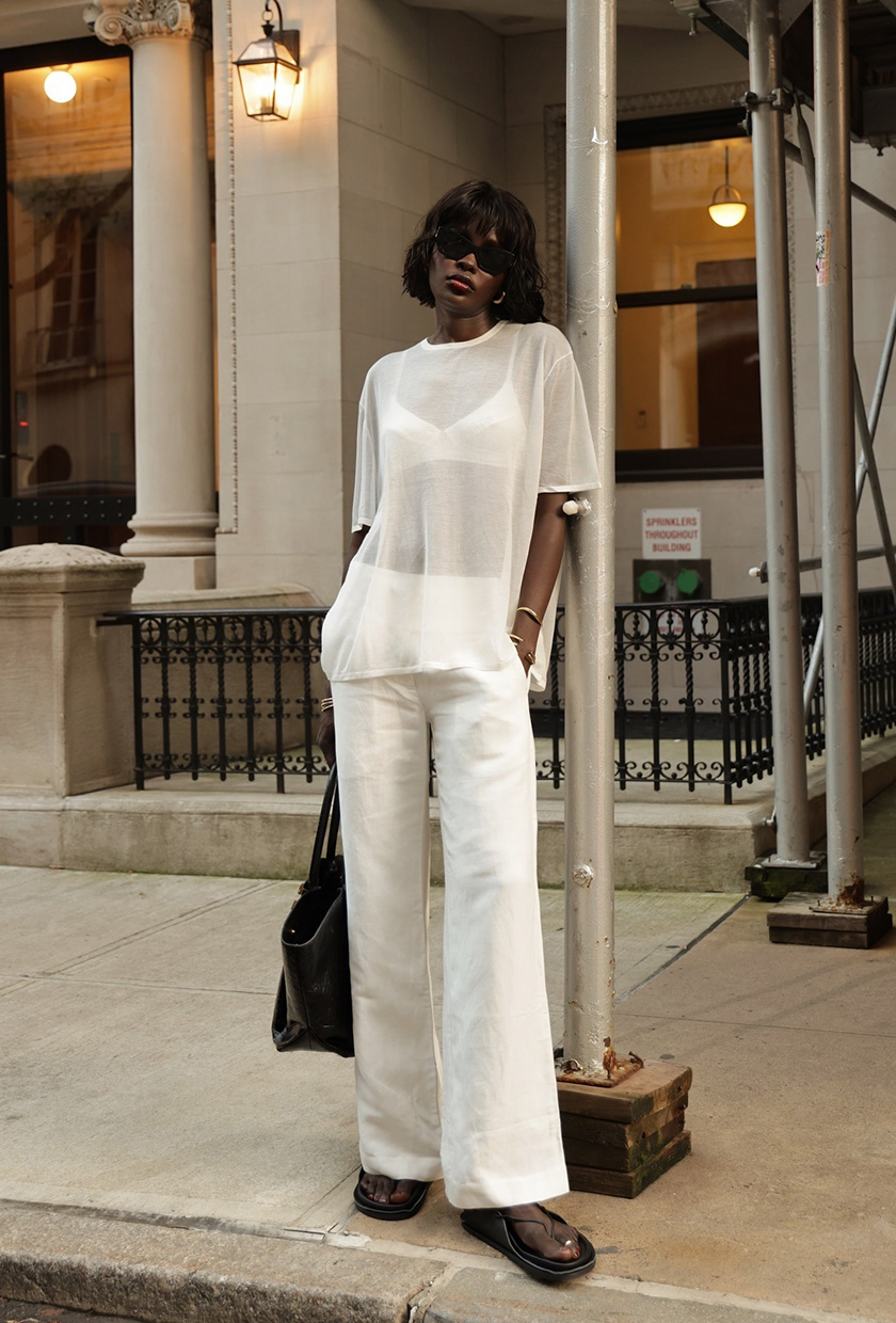 A woman standing outside on the sidewalk wearing a white oversize sheer t-shirt over a white satin bra with white linen wide-leg trousers, black flat sandals, a black tote bag, gold bangles, and black sunglasses.
