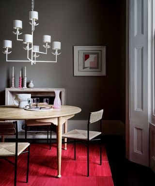 Dining room painted a dark gray-brown, light wood rounded dining table, large white chandelier with individual shades, woven dining chairs with black metal frame, traditional stone fireplace, bright red rug beneath dining table