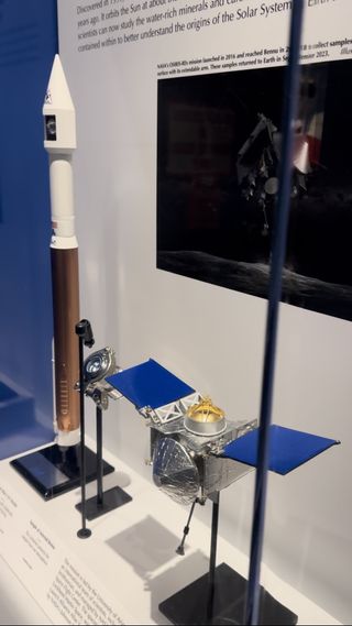 A view of the exhibit holding scale models of the OSIRIS-REx spacecraft and Atlas V rocket. In the center is the container holding the Bennu sample.