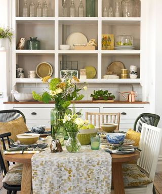 a dressed kitchen table with a white, brown and grey pattern runner, with plants on top, in front of a large white kitchen display and storage unit