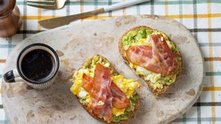 Crispy bacon on top of scrambled eggs and avocado toast with a black coffee