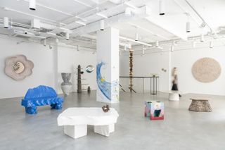 Multiple sculptural artwork including a flower-shaped mirror, a stone-carved bench and a table, a totem-like shelving unit and a series of geometric coffee tables is on view in a brightly lit gallery.