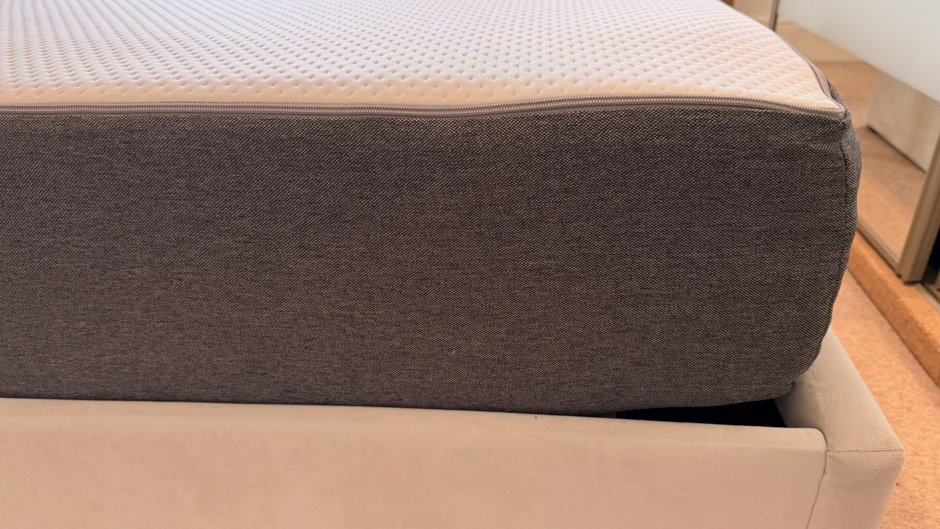 The image shows the corner of a grey and white mattress, which is placed on a grey fabric bed frame