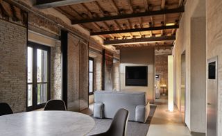 The Rooms of Rome apartment with exposed timber roof beams, exposed brick walls, grey sofa and marble dining table