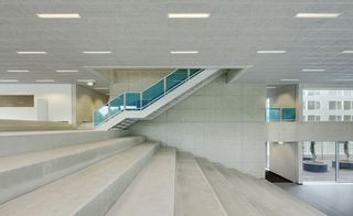 Inside image of Campus Hoogvliet, Rotterdam, white tiled walls and ceiling with strobe lights, white stone steps, white stairwell with blue transparent panels, glass fronted entrance area, grey floor, surrounding area can be seen through the ground level glass