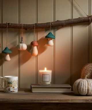 A kitchen corner with sage green paneling, a branch with mushroom decorations on it and a wooden worktop with a candle, a cup, a book and two fabric pumpkins