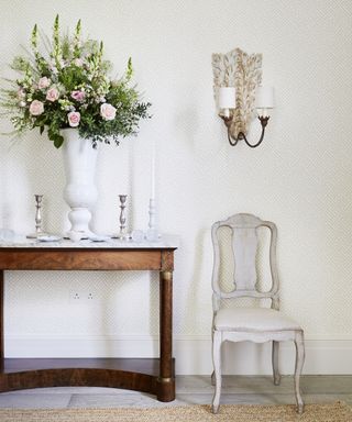 An entry table decor idea with antique wooden table, vase of pink flowers, antique chair and candle wall sconce