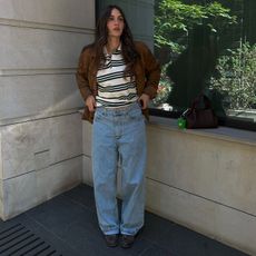 Borislava wears brown suede jacket, rugby shirt, wide jeans, and dark close-toed shoes while posing for the camera outside