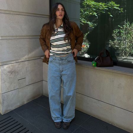 Borislava wears brown suede jacket, rugby shirt, wide jeans, and dark close-toed shoes while posing for the camera outside