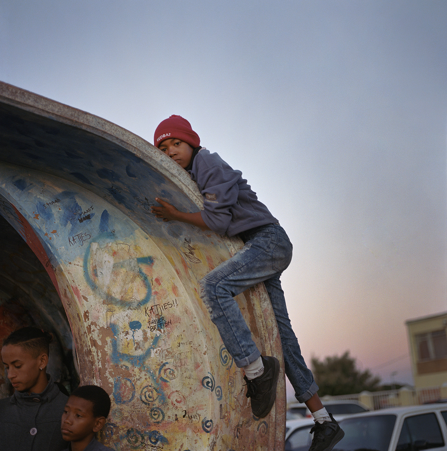 A boy in a red beanie, gray hoodie, and jeans climbs over the curved roof of a brightly graffitied structure at a bus stop during a dusky sunset, while two other boys stand below him.