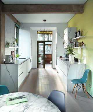 A modern white kitchen with statement wall in lime green, teal bleu chairs and marble surfaces.