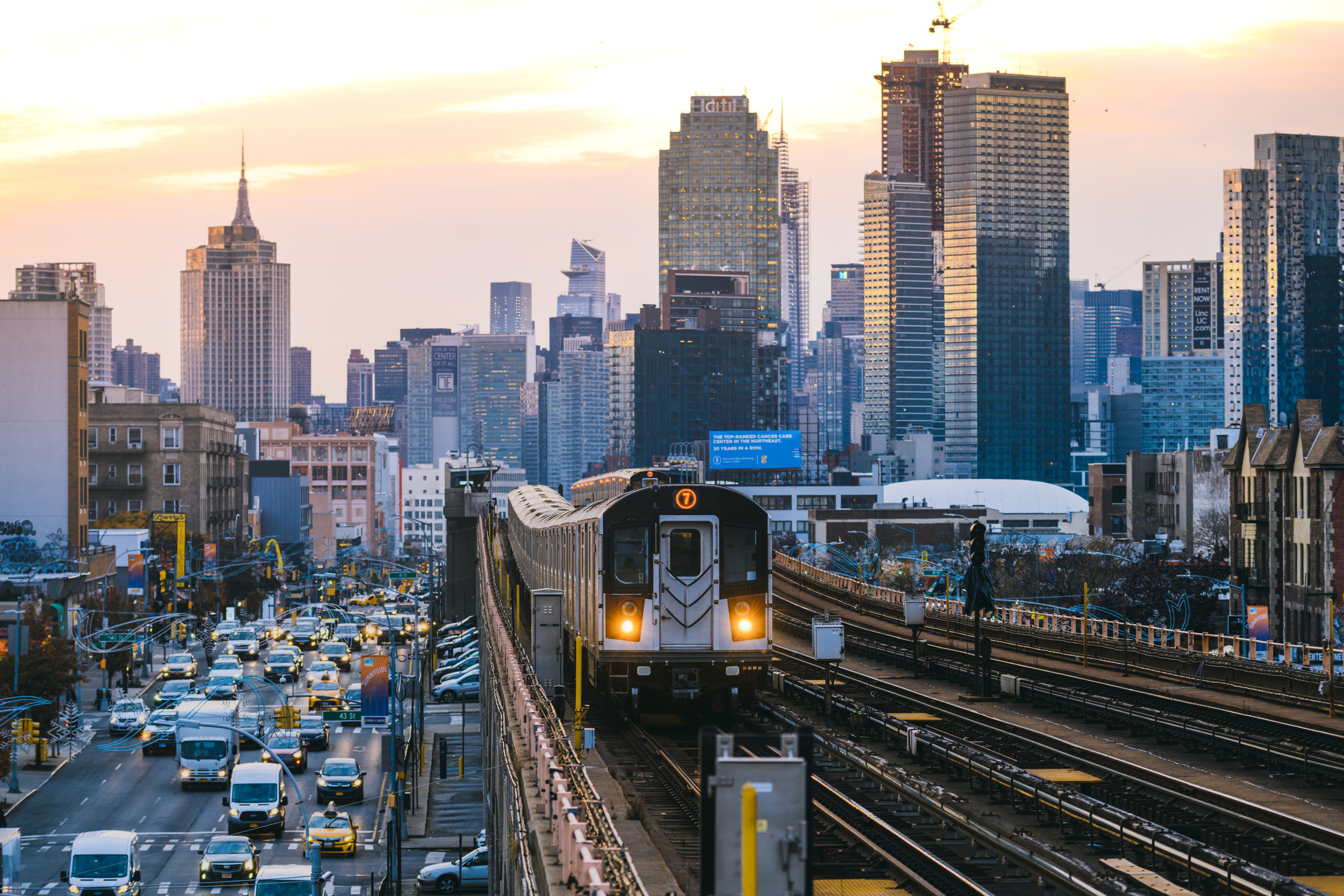 A number 7 train approaches on an elevated track in Queens, with Manhattan buildings in the background.
