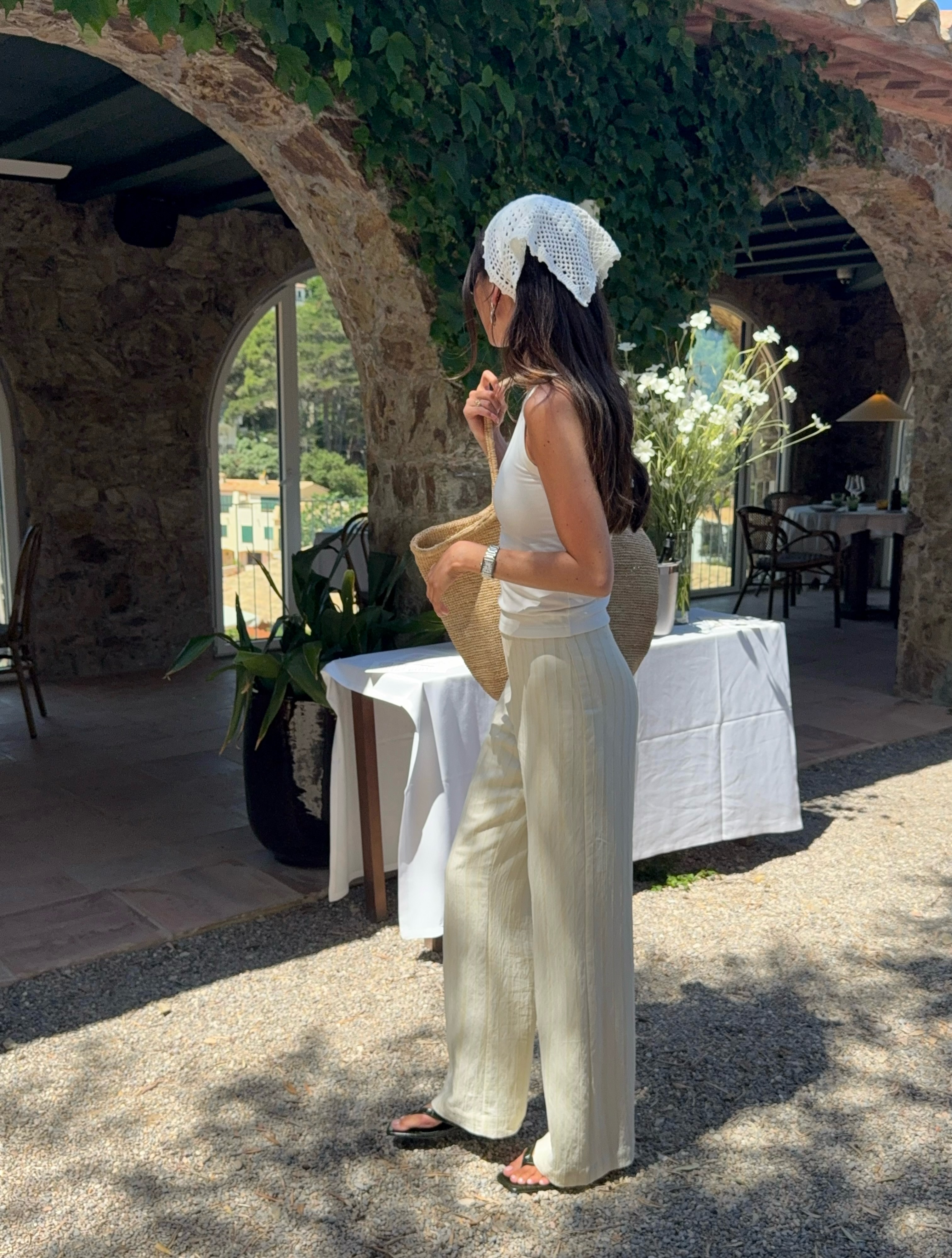 An image of an influencer wearing a crochet triangle scarf on her head, a white vest, beige trousers, black flip-flops and a basket bag.