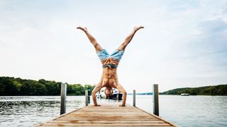 A fit man doing a handstand on a pier at a lake