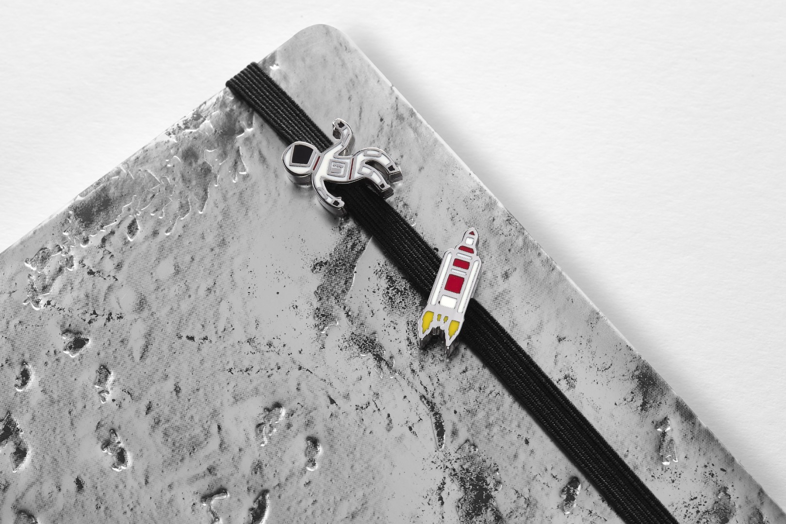Close up of two small pins attached to the black elastic closure of the NASA-inspired Limited Edition Notebook from Moleskine (silver moon surface). The top pin is a person in a white astronaut suit with red trim. The bottom pin is a small grey, white, and red simplistic rocket blasting off.
