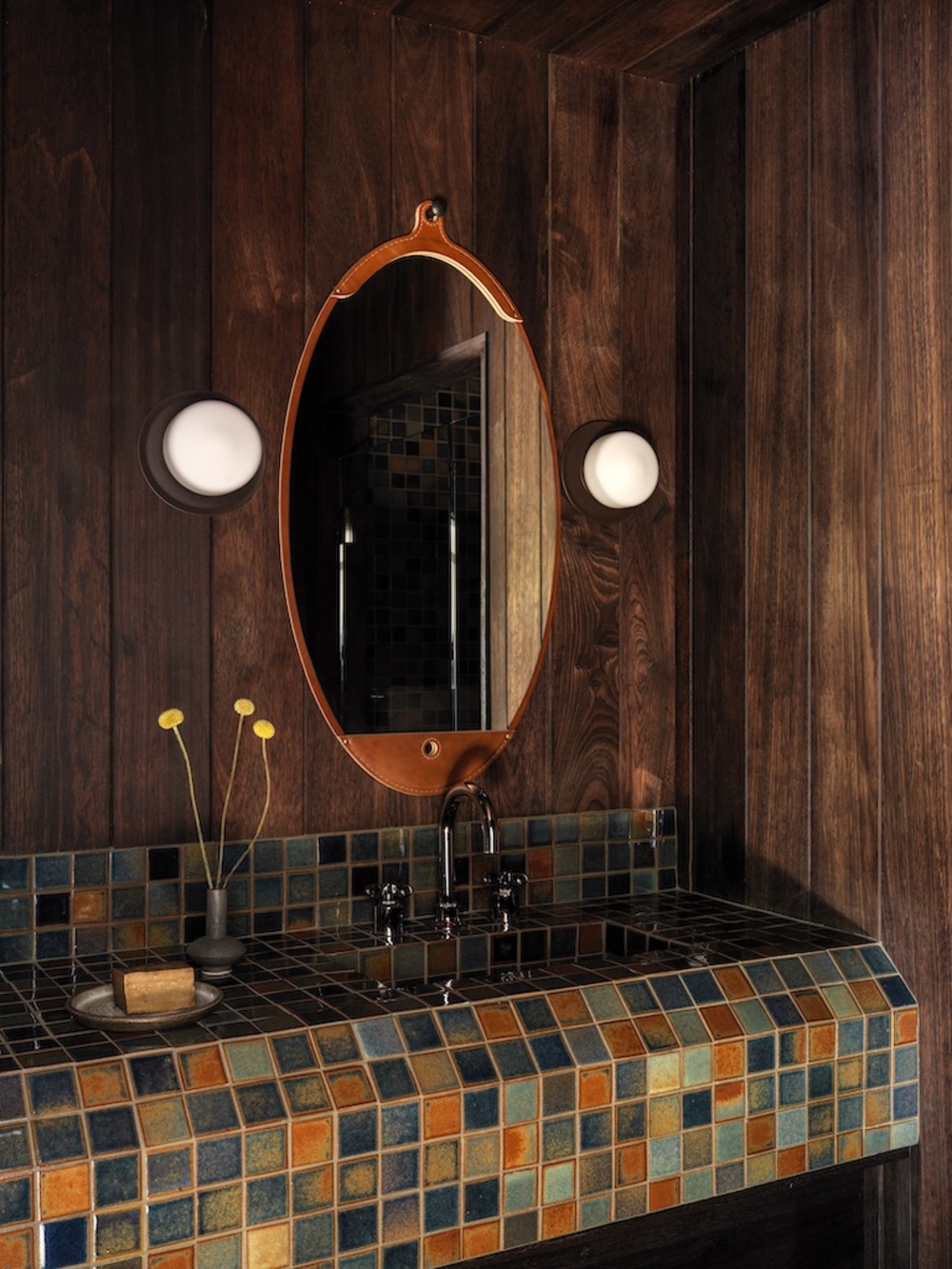 bathroom with timber walls, wall sconces on either side of an oval mirror, and colorful tiles countertop