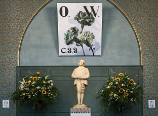 The Oscar Wilde Temple, close up of the Oscar Wilde cream statue, tall potted flowering plants, decorated walls, green arch with gold trim, O/W flower painting above the statue