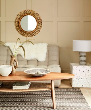 A warm beige living room with a small round ,irror on the wall above the sofa, a long, oval coffee table and a small white side table with lamp