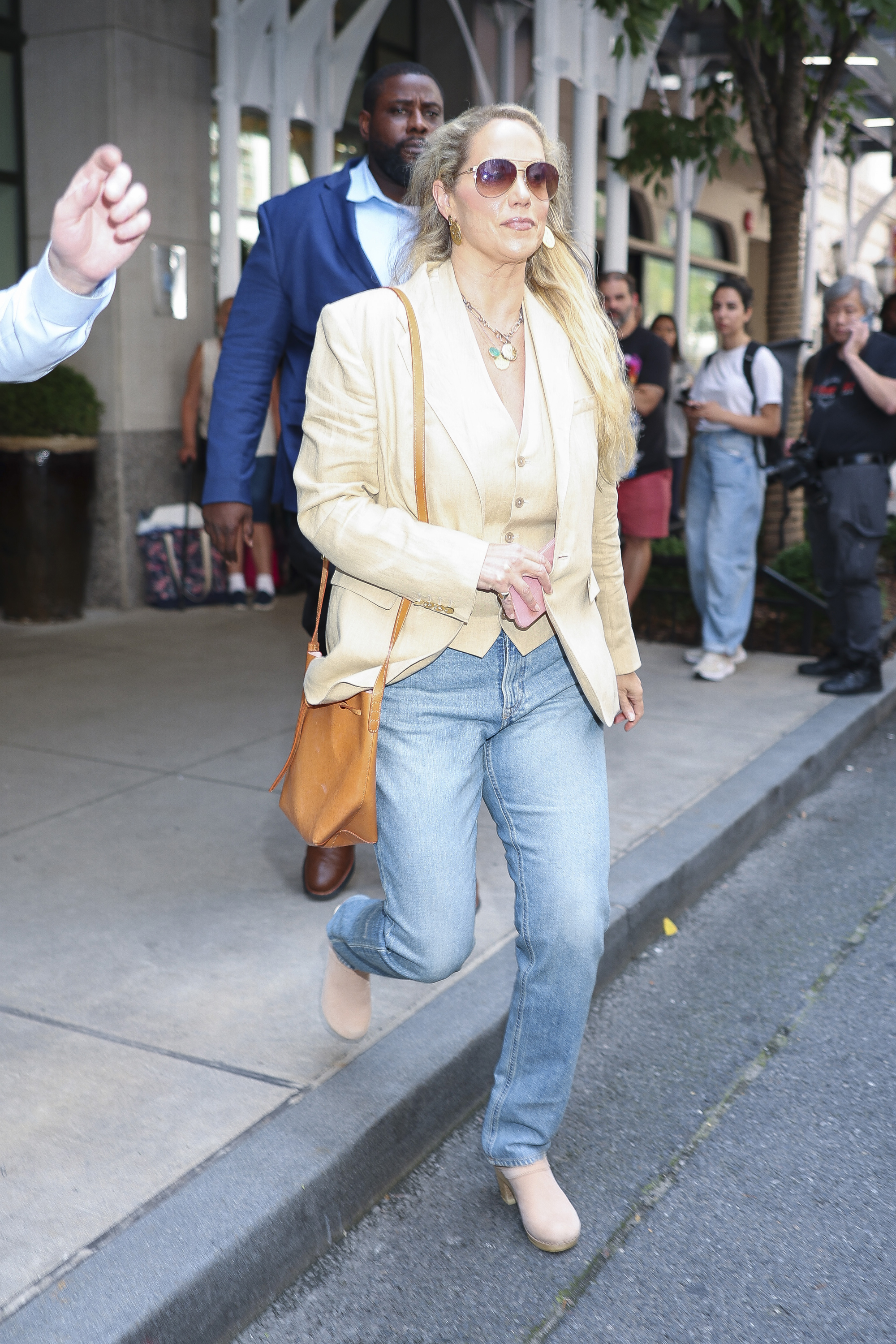 Elizabeth Berkley in NYC wearing suede clogs, a beige blazer, and a vest with light wash jeans.