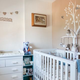 White cot in front of beige walls with tree design on, complete with wooden frames and wall garlands