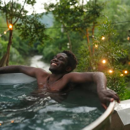 A smiling man relaxes in an outdoor tub surrounded by plants and string lights