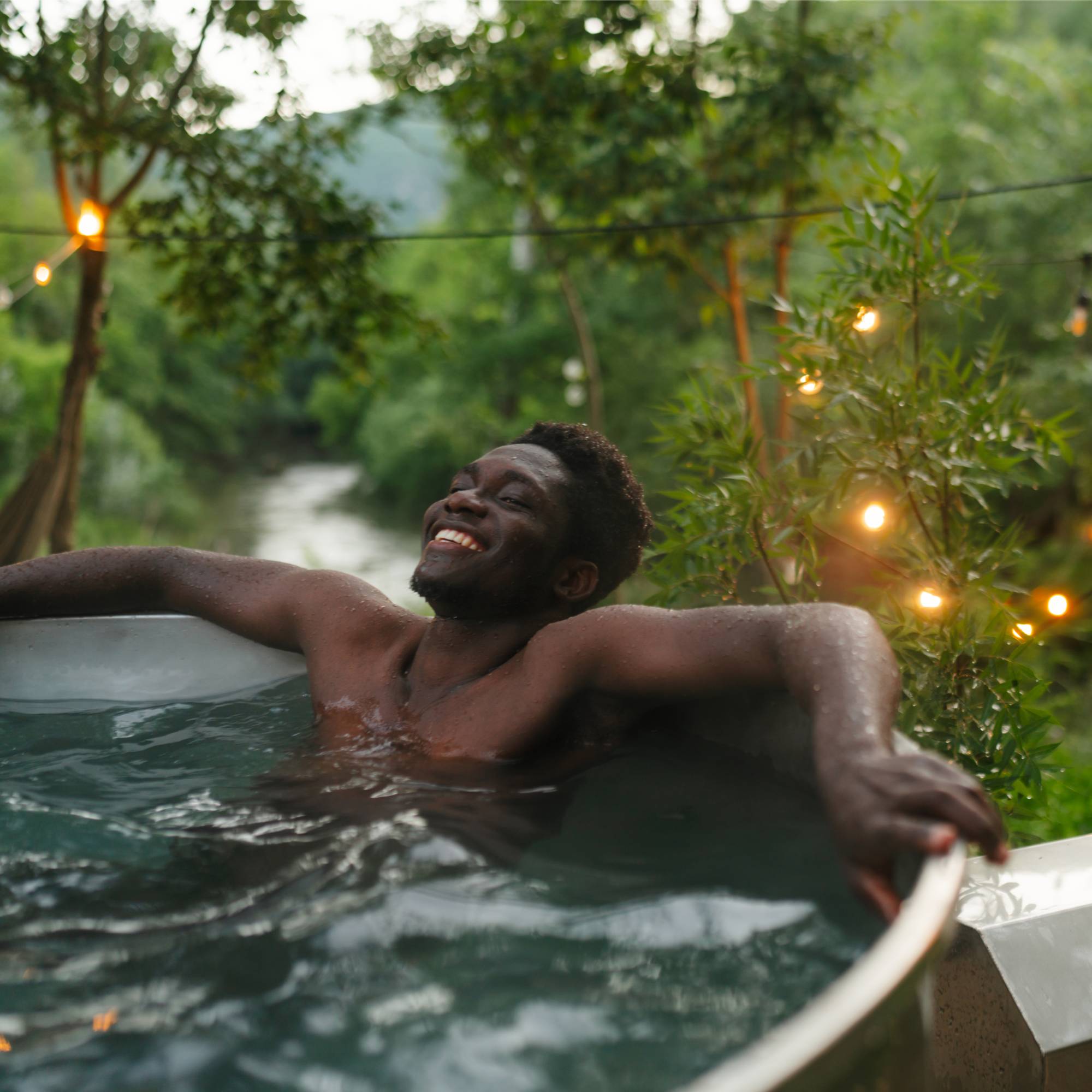 A smiling man relaxes in an outdoor tub surrounded by plants and string lights