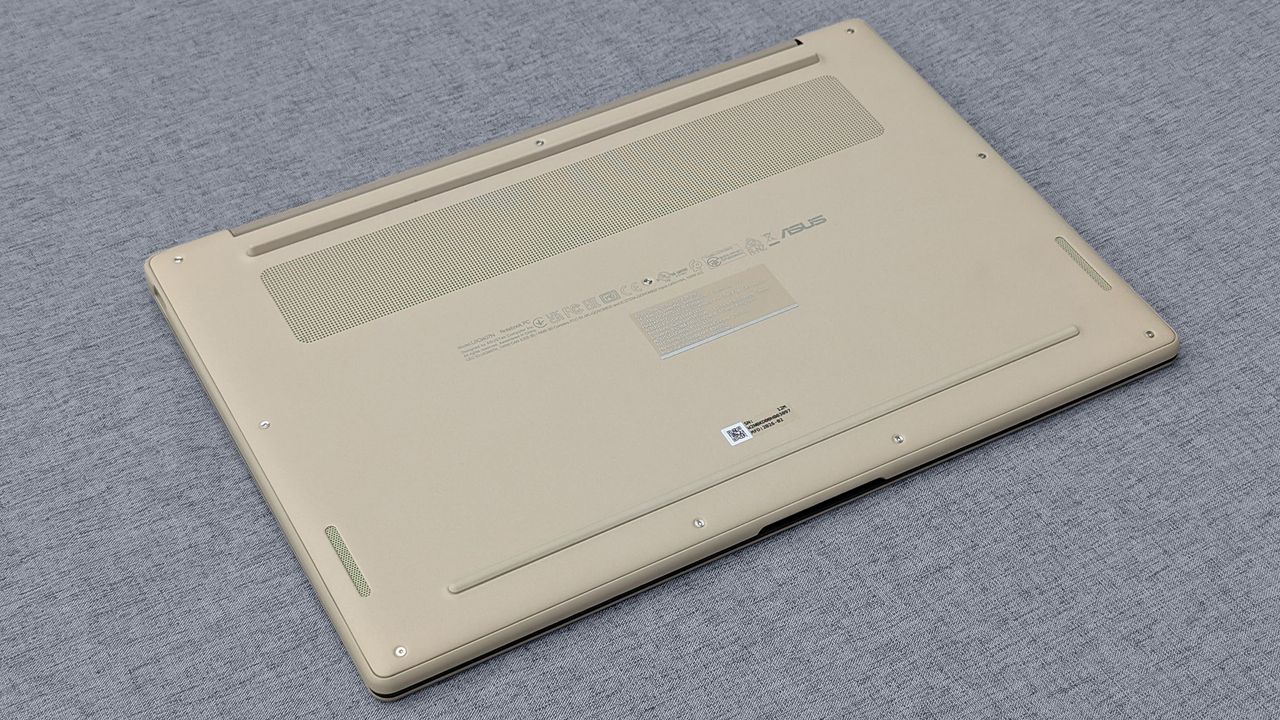 Underside of beige ASUS Zenbook A14 with ventilation grilles, rubber feet, screws, and regulatory markings on gray fabric.