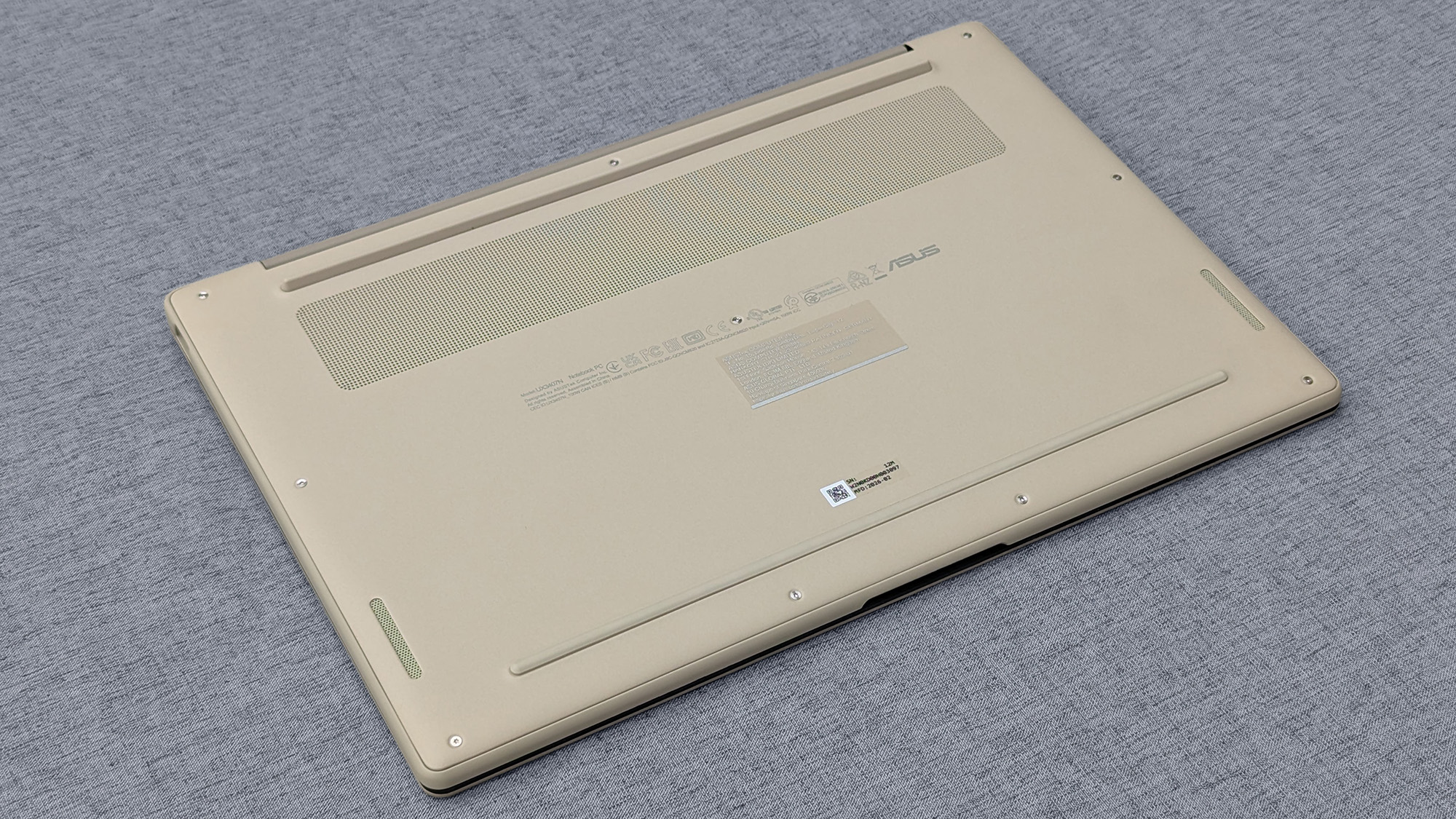 Underside of beige ASUS Zenbook A14 with ventilation grilles, rubber feet, screws, and regulatory markings on gray fabric.