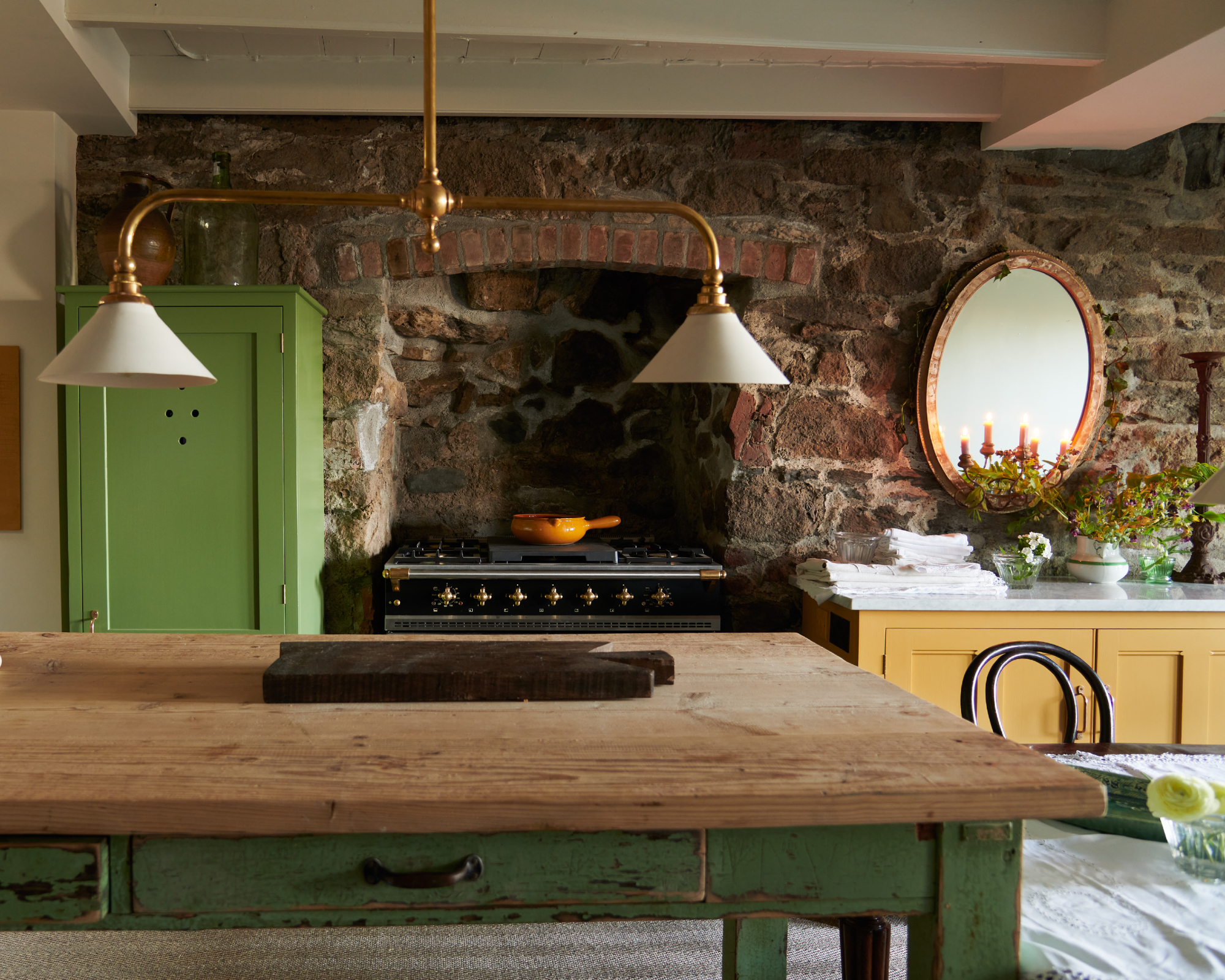 green and yellow farmhouse style kitchen with freestanding furniture, enamel lights, stove, original bricks, napkins/tablecloth, mirror