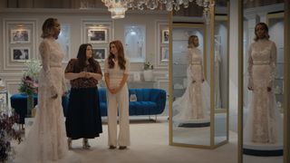 Helen trying on a wedding dress at a bridal shop and looking at herself in the mirror. Dana Sue and Maddie are standing next to her and looking at each other.