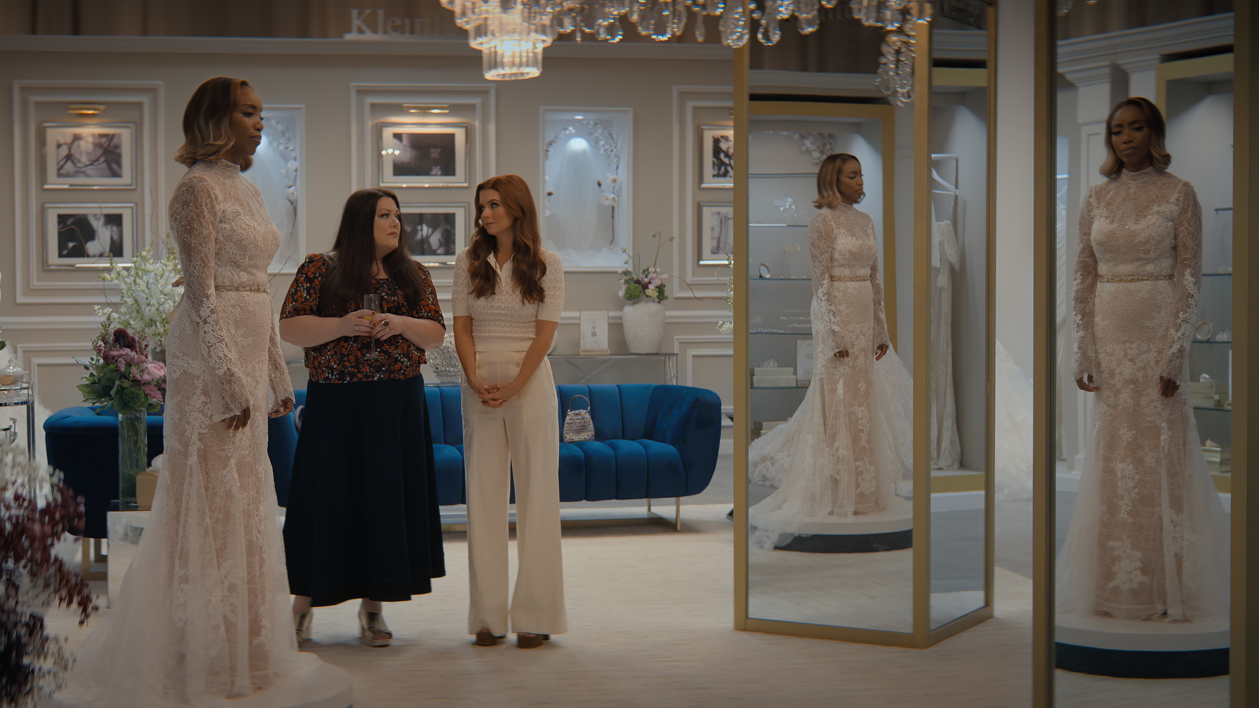 Helen trying on a wedding dress at a bridal shop and looking at herself in the mirror. Dana Sue and Maddie are standing next to her and looking at each other.