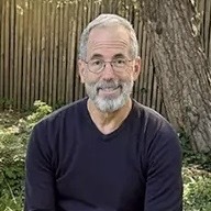 A middle aged man with grey hair and beard wears glasses and v-neck long-sleeve shirt