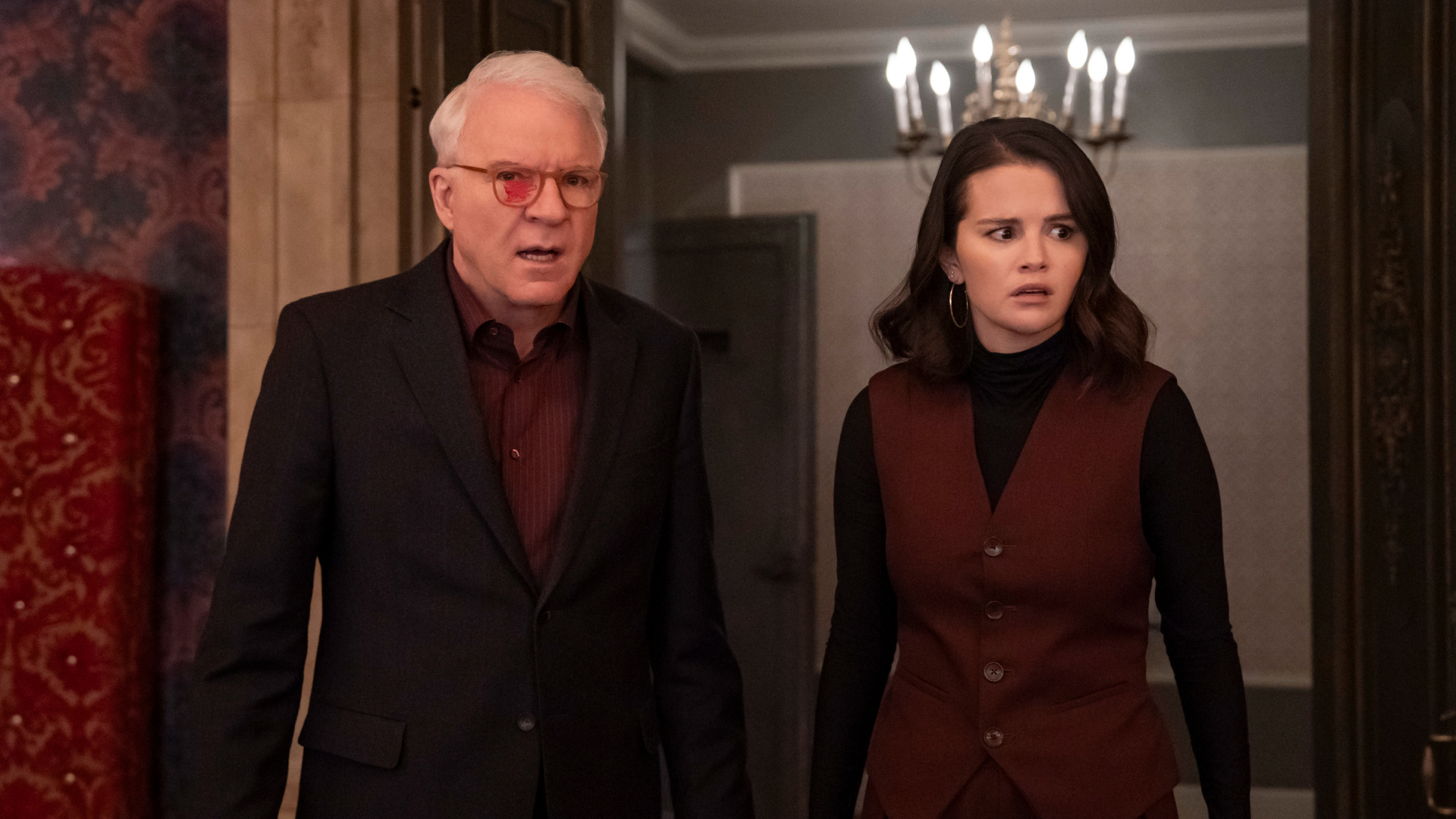 A man (Steve Martin as Charles) wearing glasses with red marker on the left lens stands with a woman (Selena Gomez as Mabel) in a ballroom doorway with a chandelier behind them, in 'Only Murders in the Building.'