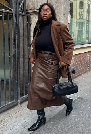 A black turtleneck outfit is shown in a photo of a woman standing on the sidewalk wearing a brown suede blazer over a black turtleneck with a black skinny belt and a brown leather skirt, black knee-high boots and a black shoulder bag