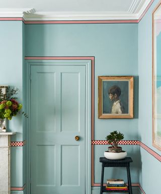 Painted blue hallway with blue painted door, light wooden flooring, red stripes and checkered boards around trims. Artwork beside door, black stool with books and plants.