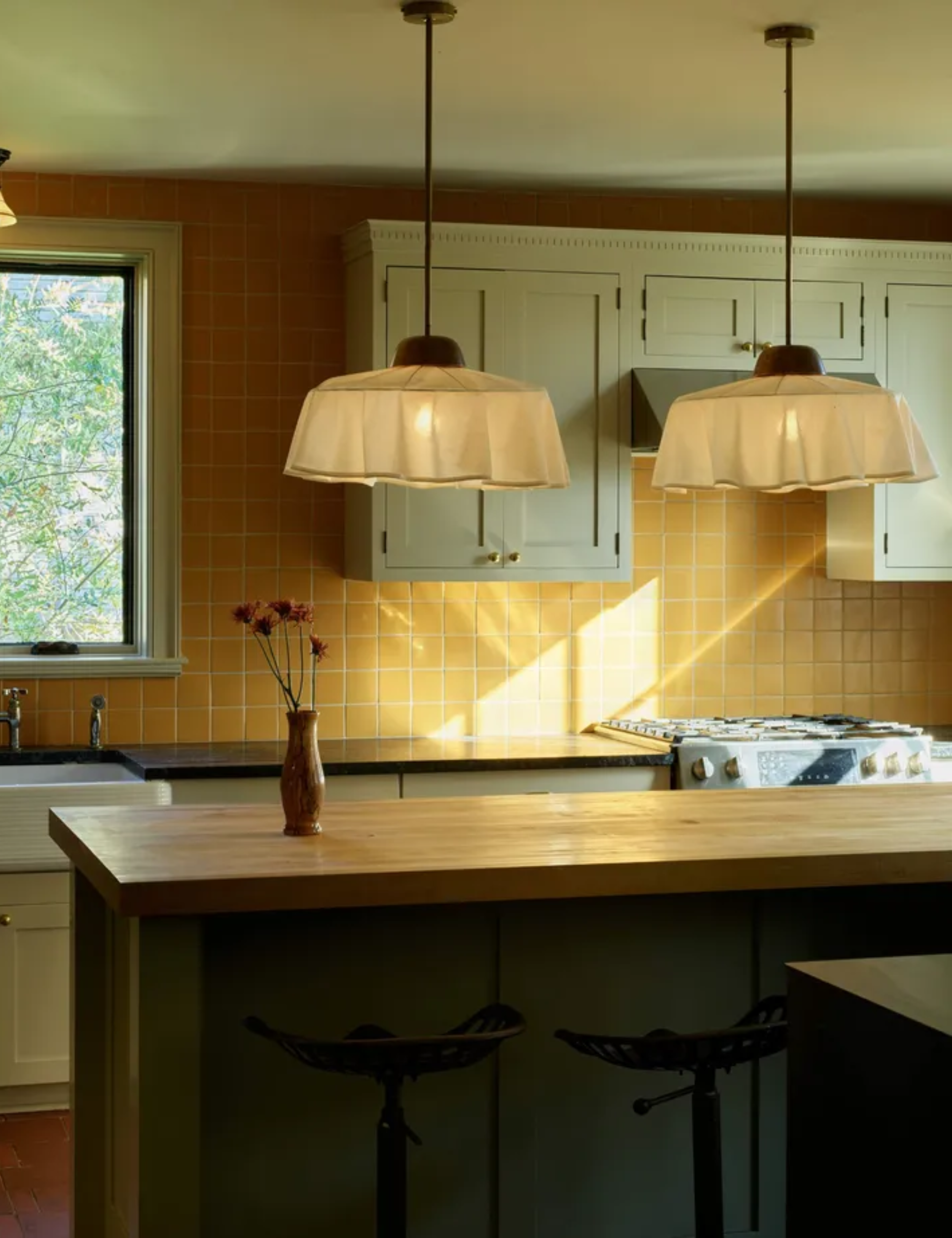 Kitchen with light wood butcher block counter, two white ruffled pendant lights, off-white upper cabinets, and a backsplash made of small, square orange/yellow tiles