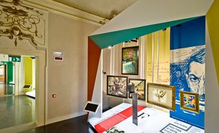 Exhibition neutral room with black gloss floor, Italian futurism display, colourful framed canopy above framed artwork, information display, doorway with gold detail above the opening, view of the gallery beyond