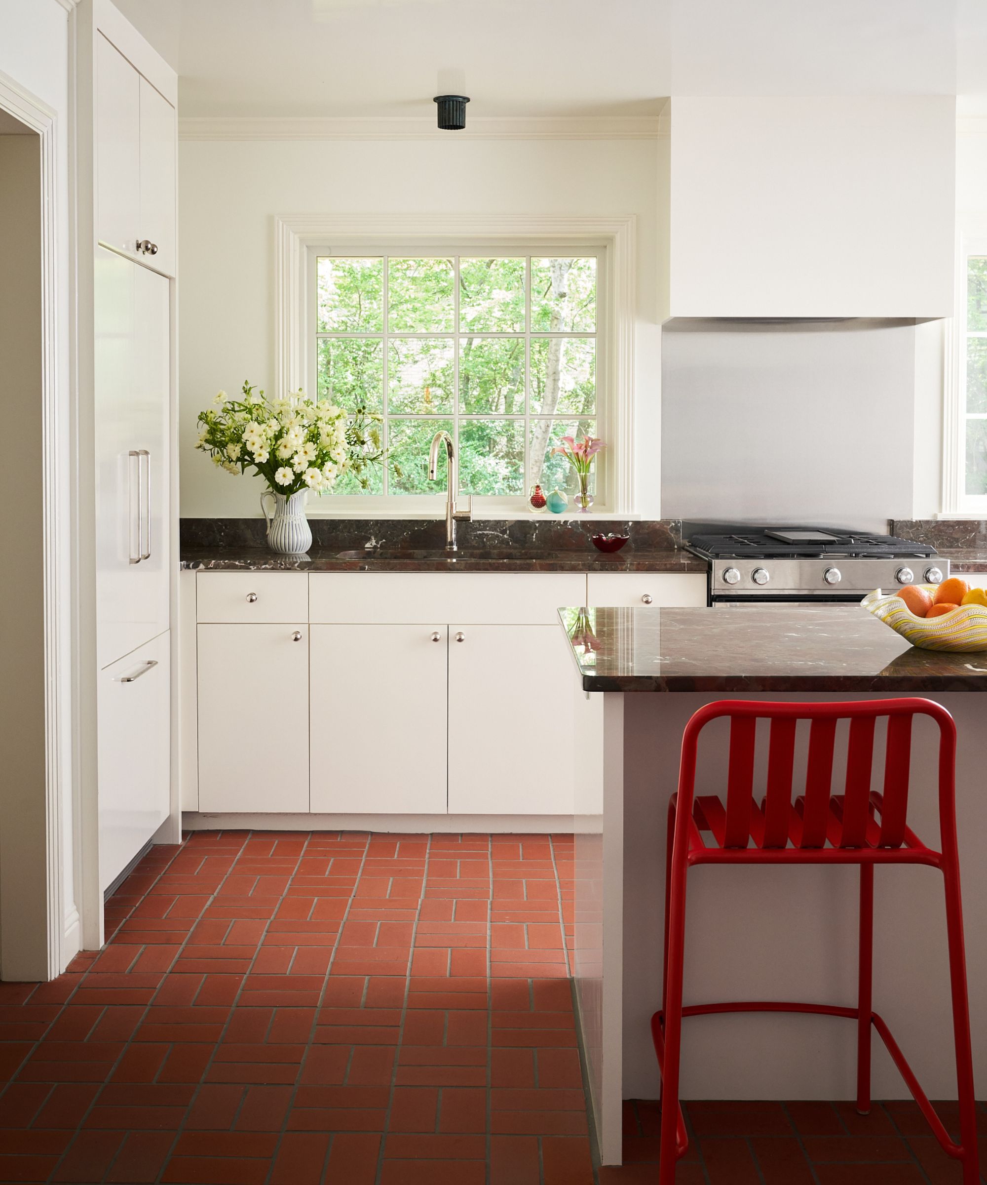 a sleek white kitchen with dark red marble counters, plaster cooker hood, brick paver tiled floor and red kitchen island stools
