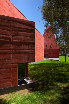 A reddish concrete building on a green field beside tall trees