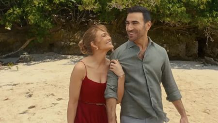 JLo wearing a red dress and hanging onto Brett Goldstein's arms on a beach in Office Romance.