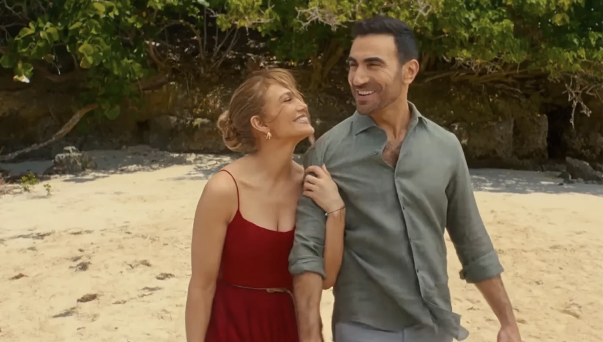 JLo wearing a red dress and hanging onto Brett Goldstein's arms on a beach in Office Romance.