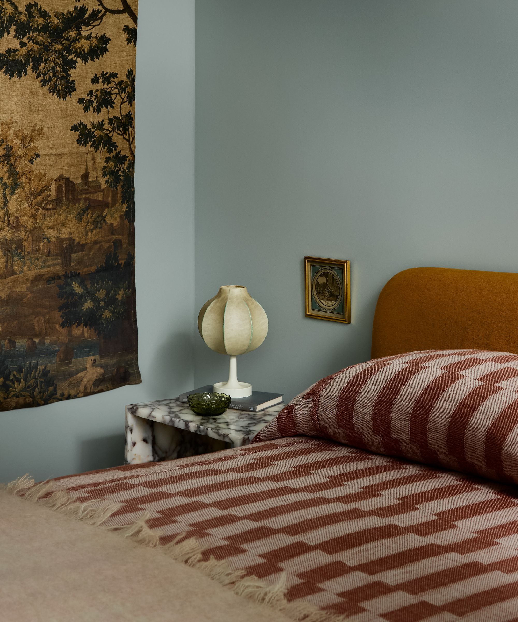 a light blue painted bedroom with a tapestry wall hanging, marigold yellow headboard, rust red printed bedding and a marble side table