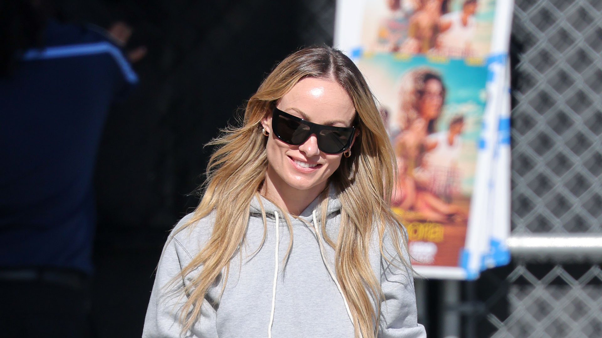 Olivia Wilde wearing a gray hoodie, black trousers, black pointed-toe shoes, and black sunglasses