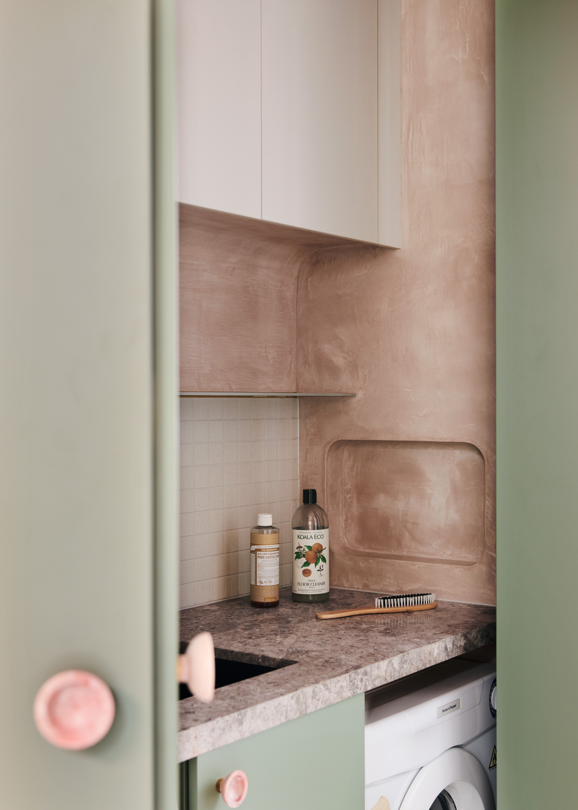 A laundry room with a blush limestone wall, sage green cabinetry, pink hardware, a marble countertop, and a white washing machine