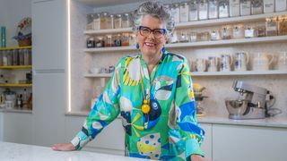 Prue in her kitchen smiling for the camera and wearing a bright top and necklace.