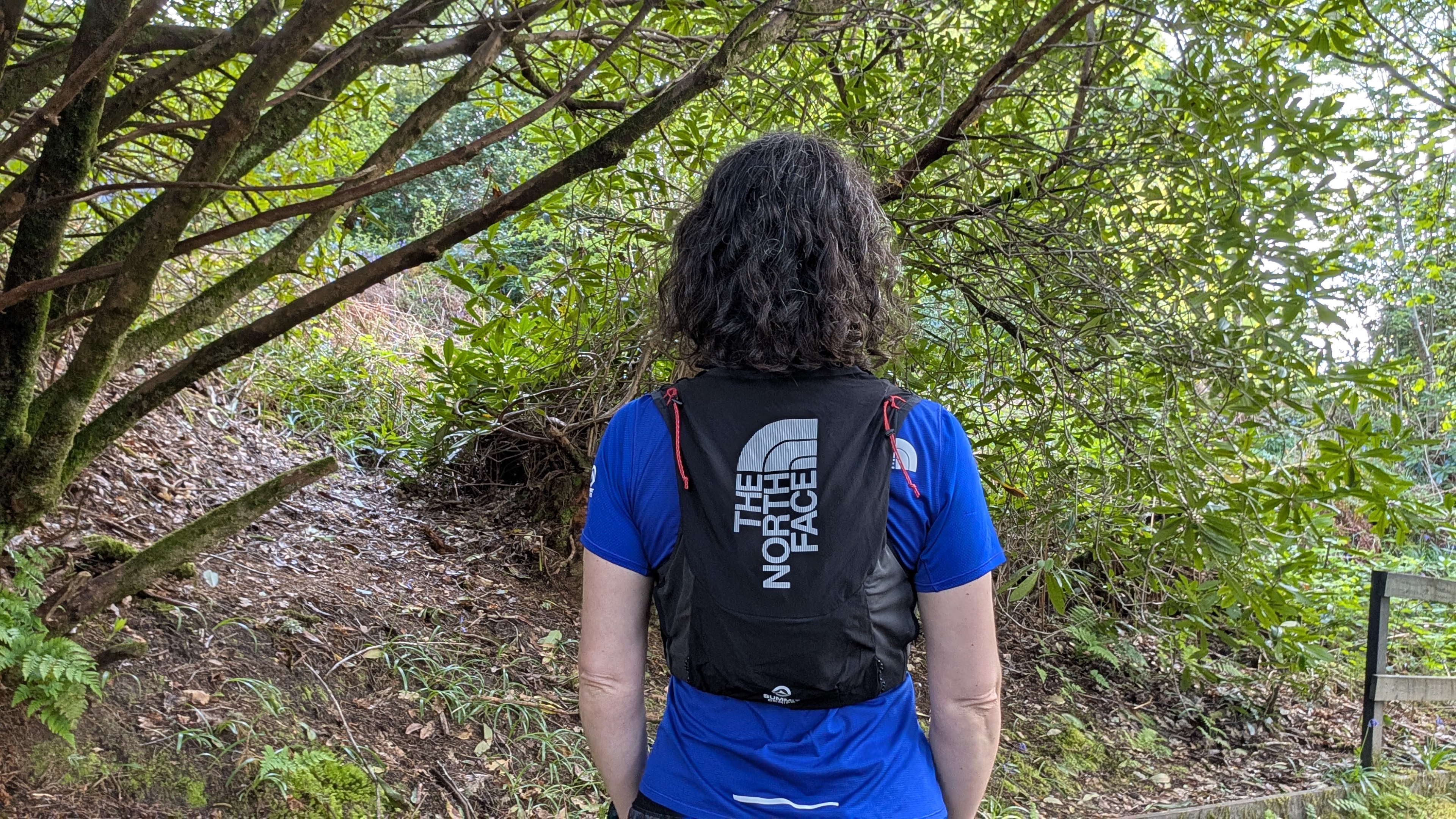 A woman trail runner from behind wearing a blue shirt and black vest with The North Face logo in white