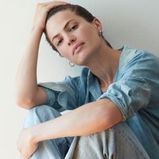 Cameron Russell sits down against a plain wall wearing a denim shirt with the sleeves rolled up and gray pants