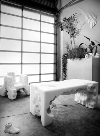 Black and white photograph of Daniel Arsham furniture, including a white resin table and chair whose design looks like eroded rock
