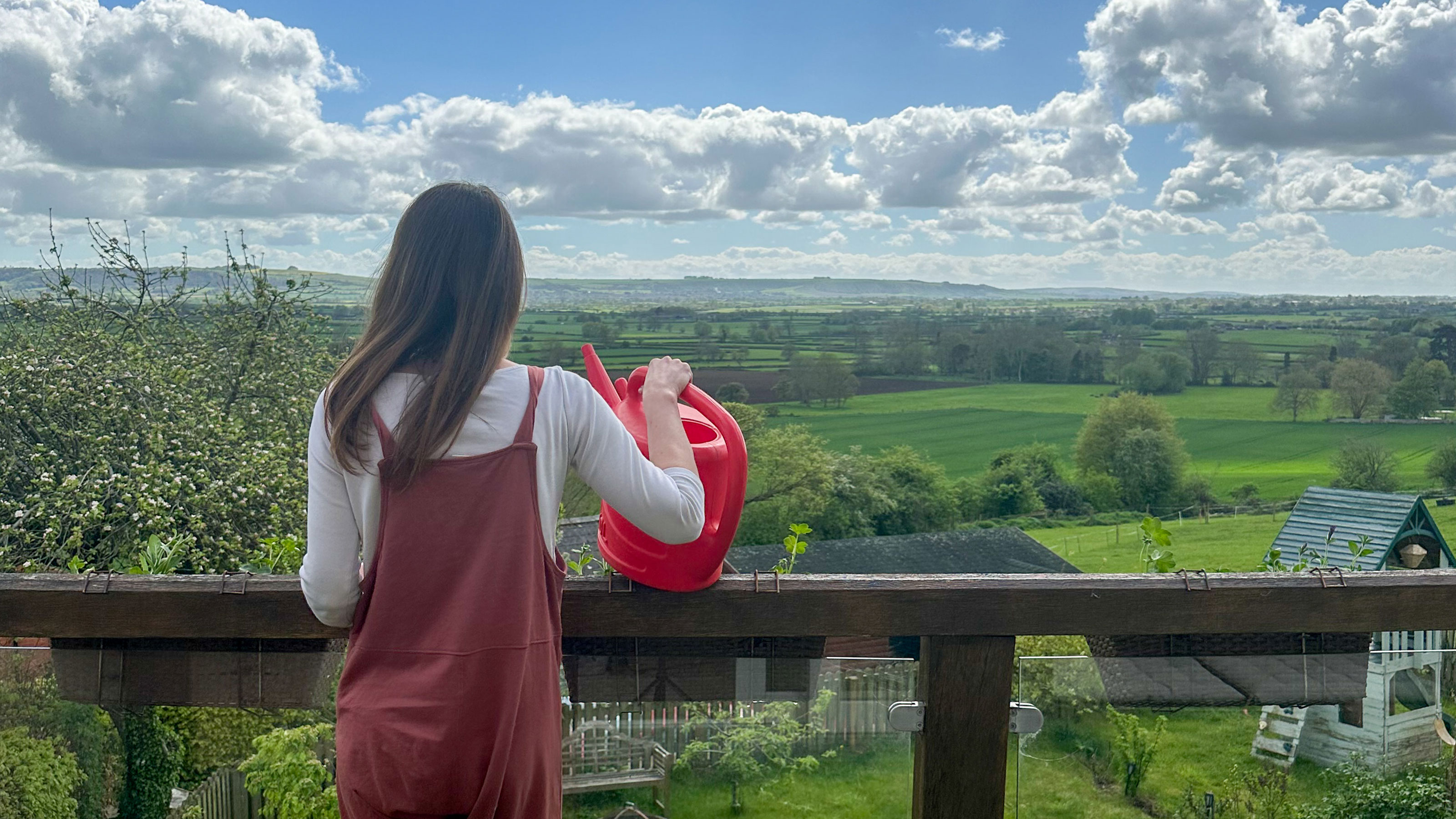 Woman holding watering can standing on balcony overlooking garden and fields