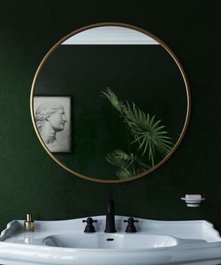 A dark green bathroom with a circular gold mirror reflecting a wall art print and plant, and a white basin with black faucets underneath it
